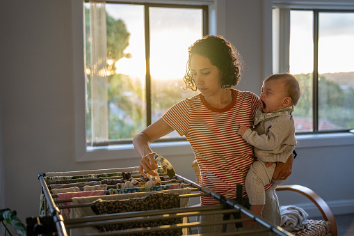 Loving stay at home mother of Pacific Islander and Asian descent holds her Eurasian 9 month old baby son on her hip as she places clean clothing on a drying rack set up in the living room at home. Golden sunlight is streaming through the window in the background.
