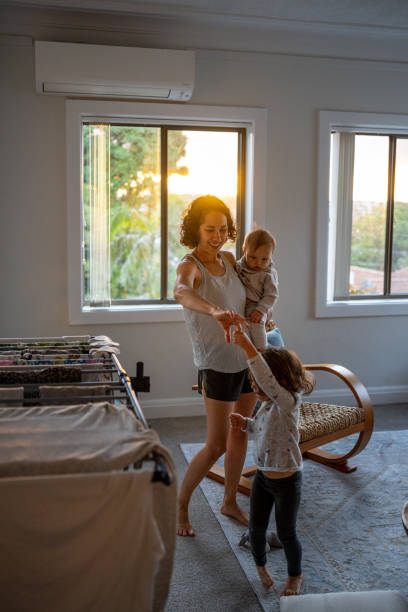 Mother holds baby son and dances with preschool age daughter Loving Eurasian mother holds her 9 month old son on her hip while dancing with her 3 year old daughter in the living room at home during sunset. baby boy dancing with his mommy stock pictures, royalty-free photos & images