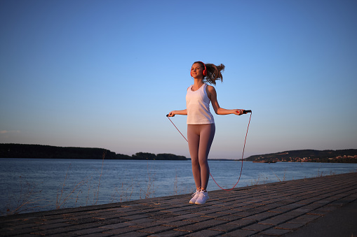 Beautiful young girl in her 20s with blonde hair by the river on a fitness path exercising with jumping rope while listening to music and enjoying the sunset. Photo taken in the golden hour