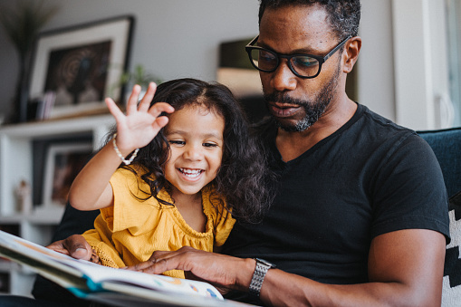 A loving father of African descent sits on the couch in the living room of his home and reads a picture book to his preschool age daughter, who is sitting on his lap smiling.