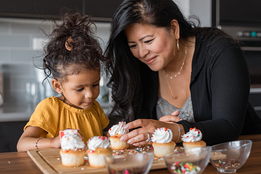 A mother of Asian ethnicity and her preschool age daughter of mixed race decorates and frosts cupcakes together, as they get ready for a family birthday celebration at home.