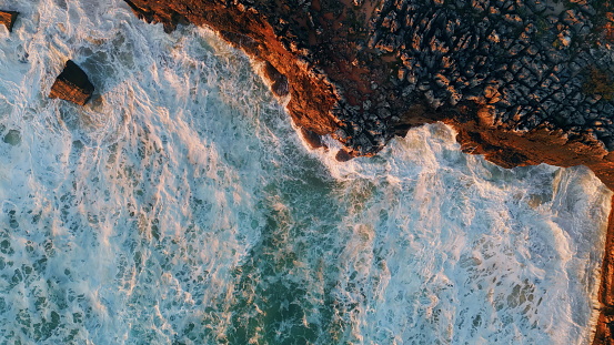 Top view foamy sea splashing under coastal cliffs in super slow motion. Picturesque ocean waves foaming bubbling at rocky seashore. Drone shot powerful water washing on stone coast. Marine scenery.