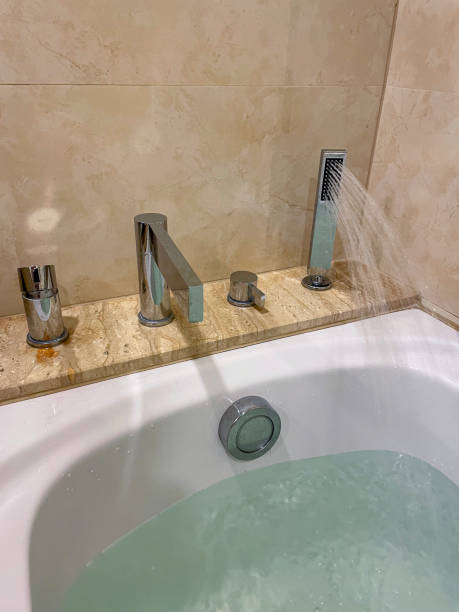 Stock photo showing a modern white bathroom suite with a bath featuring an integrated shower head and chrome mixer tap being used to fill the tub with water. The bathroom has been tiled with stone wall tiles. imagen de suite de baño blanca, rociado de agua desde el cabezal de ducha integrado, relleno moderno, bañera blanca, grifo mezclador cromado, fila de controles cromados, perilla emergente de control de residuos, azulejos de pared de piedra