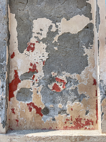 Stock photo showing close-up view of red peeling masonry paint layers on damp grey concrete wall with flaking plaster.