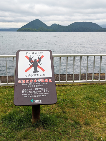 Toyako-cho, Japan - June 9, 2023: A Ministry of the Environment warning sign states not to remove live crayfish from this habitat. Background shows the Nakajima Island group in the caldera lake of Shikotsu-Toya National Park. Spring morning by the Lakeside Path.