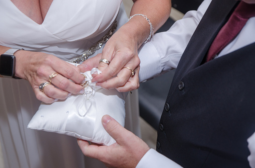 Close-up on wedding rings on a white cushion held by groom. Bride is untying the knots that holds the rings.