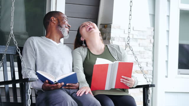 Multiracial couple on porch swing reading books, laughing