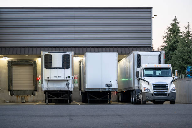 istock Day cab big rig semi truck with refrigerator semi trailer unloading cargo standing in warehouse receiving dock gate with another semi trailers at twilight time 1621982432