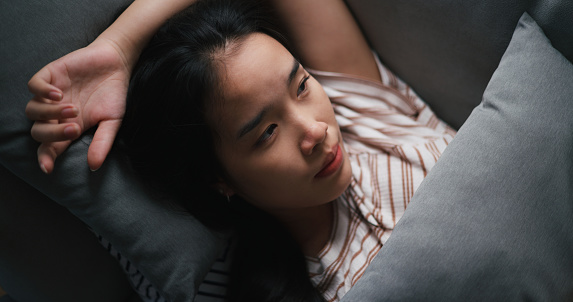 Portrait of Pensive depressed young Asian woman lying on sofa at home looking at ceiling thinking pondering, feeling sad and frustrated, having life problems, Mental distress and people concepts.
