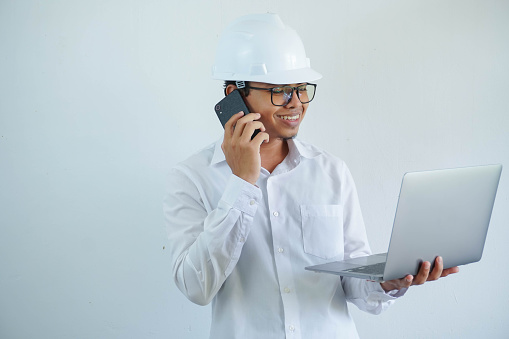 young Asian male engineer wearing white hard hat holding laptop and on call for construction work isolated on white background, copy space.