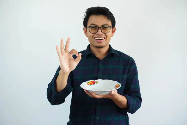 young asian man smiling happy and give OK sign with hand while showing empty dinner plate isolated on white background.