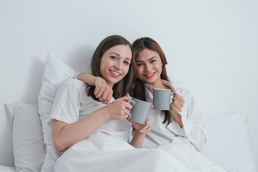 Two Asian cheerful happy female girlfriends having a coffee sitting on the bed. Concept lifestyle lesbian couple.