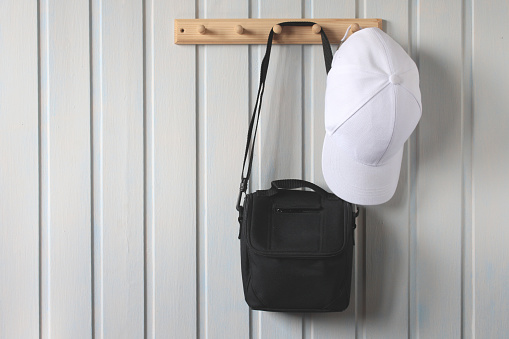 camera bag and a baseball cap hang on a white wall on a wooden hanger.