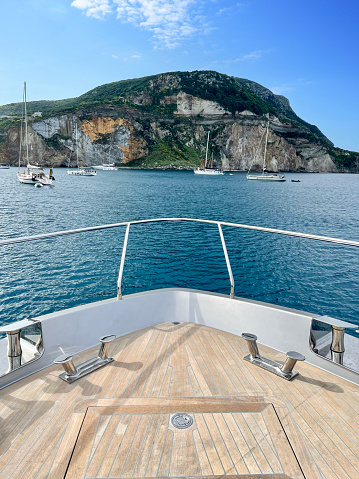 The bow of a luxury yacht approaching the far side of Ponza island in the daytime, which has sailboats anchored near it.