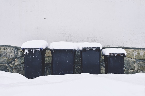 A winter scene with a collection of garbage bins lined up in front of a snow-covered wall of stone