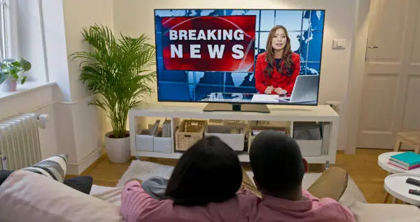 Rear view of couple cuddling and watching newscaster presenting breaking news on TV while sitting on sofa in living room.