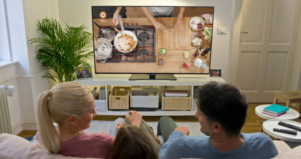 Family watching TV at home Rear view of family watching cooking show on TV while sitting on sofa at home. television show stock pictures, royalty-free photos & images