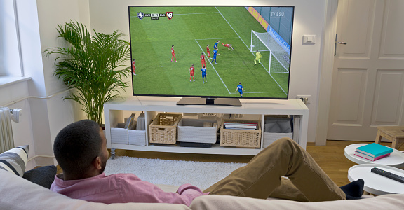 Man watching football match on TV while relaxing on sofa in living room.