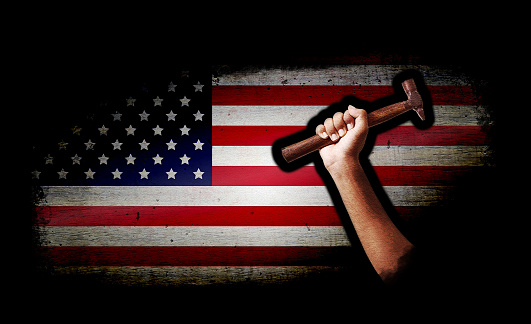 Messy grungy black colour vignette on wall with grunge texture with one hand emerging from it holding a work tool hammer with American flag in backdrop. Apt for use as posters, backdrops, banners, greeting cards for USA Labor day, May day.
