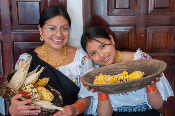 Photo of two smiling indigenous people from otavalo in traditional dress showing a basket with the harvest of ears of corn