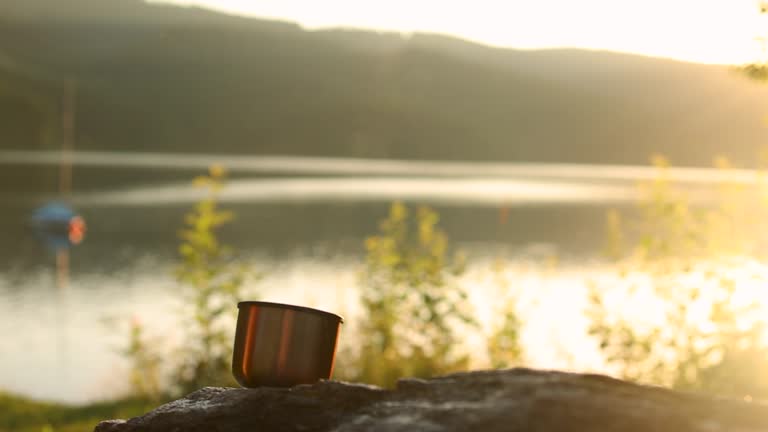 A man's hand pours tea from a thermos into a mug at sunset