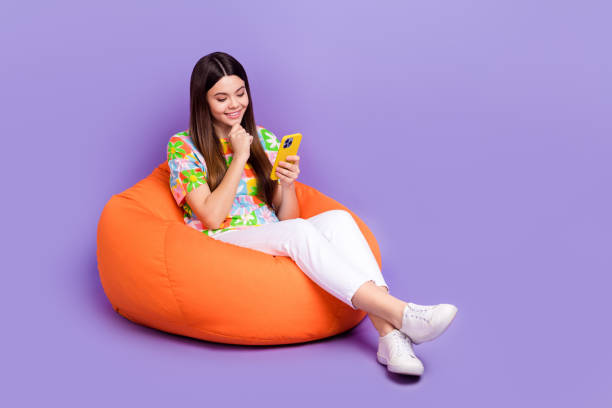 Full body photo of smiling teen girl touch chin using smartphone good mood texting friends lying pouf isolated on violet color background stock photo