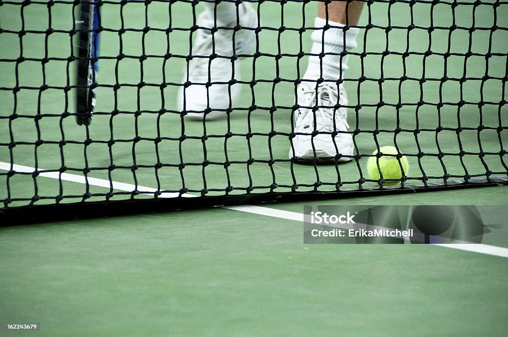 Feet approaching a ball on the tennis court A man approaches a tennis net to pick up a ball that is lying on the ground of an indoor court. Tennis Stock Photo