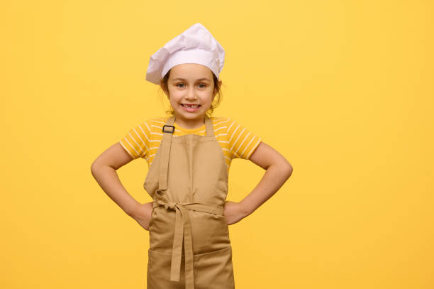 Mischievous child girl dressed as confectioner in apron and white chef's cap, putting hands on waist, looking at camera stock photo
