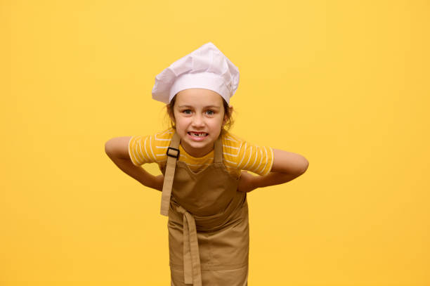 istock Mischievous little girl in chef's hat and apron, grimacing, making faces, posing with hands on waist, isolated on yellow 1622440000
