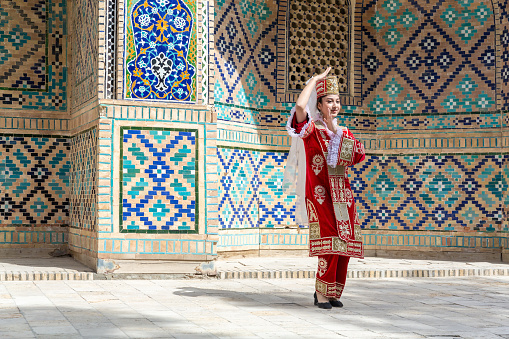 Young Uzbek woman dancer in red national clothes, historical building at background. Folk show, public space. Travelling, national culture, entertainment concept. April 20, 2023 - Bukhara, Uzbekistan