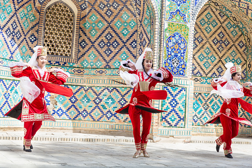 Three Uzbek female folk dancers in red national clothes dabce in center of Buxoro near mediaval medrese. Art, entertainment and national culture concept. April 20, 2023 - Bukhara, Uzbekistan