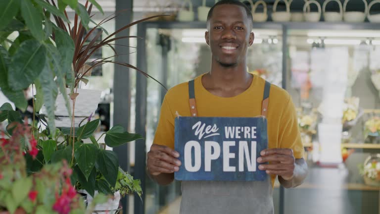 Portrait of young African American man entrepreneur holding Yes we are open sign in new flower store