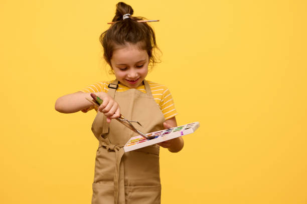 웃고 있는 어린 소녀 예술가가 수채화 물감에 붓을 담그고 고립된 노란색 배경에 담근다. 광고 공간 복사 - palette knife painting 뉴스 사진 이미지