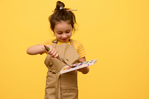 Smiling little child girl artist, schoolgirl dressed as an artist in beige apron, learning painting art, dipping paintbrush into a palette with watercolor paints, isolated yellow backdrop. Copy space