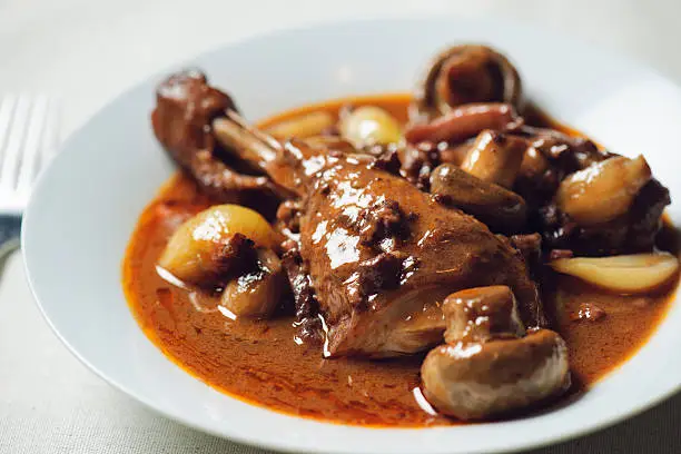 Coq au vin -- chicken stew -- on a fancy white plate with a fork on the side.