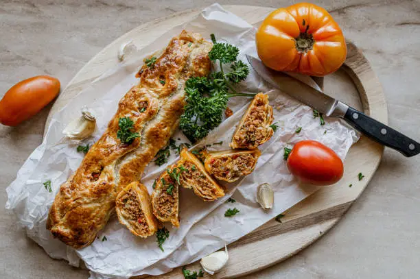 Delicious homemade puff pastry rolls or pockets. Filled with tasty turkish minced meat. Served whole and sliced on a light wooden cutting board isolated on a table. Top view with copy space