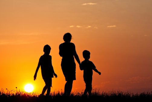 A silhouette of a mother walking with her children at sunset. Family. Leisure. Walking. Rurual setting. Panorama. Themes include health, vitality, families, mother, parenting, exercise, evening, care, together, fun, evening, meadow, rural, prairie, scenic, three people, boys, siblings, sons, people, children, group, single mother, and fitness. Back view. People are unrecognizable. 