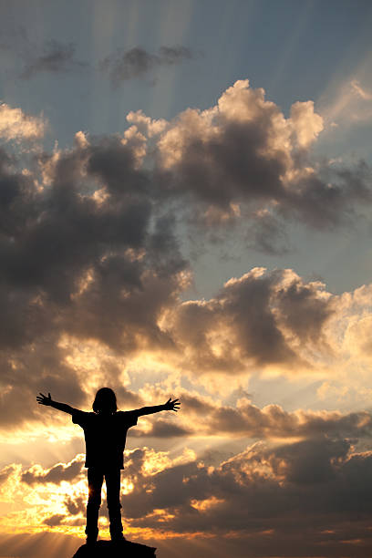 Happy Child in Worship A happy child raising his arms to the sky at sunset. Vertical colour image. Copyspace. Worship concept. Silhouette. Natural God beams in the sky. religion sunbeam one person children only stock pictures, royalty-free photos & images