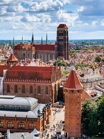 Aerial view of the Gdansk Old town with St. Mary’s Church and Baszta Jacek tower. The image is taken from above, showing the rooftops and streets of the historic city center. The sky is blue with white clouds and there are people walking on the pavement below.