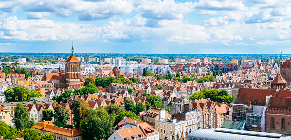 A panoramic view of Gdansk old town centre. The church is Kościół pw. św. Jana (St. John’s Church) is on the background. The sky is blue with white clouds and the city is surrounded by trees. The image is taken from a high vantage point, looking down on the city.