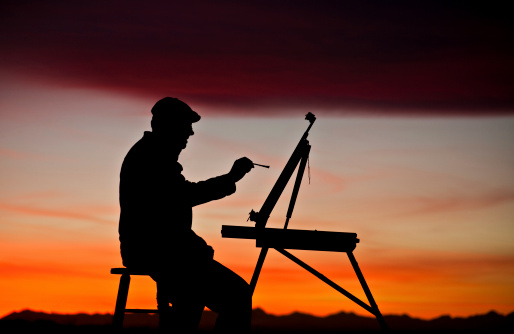 A silhouette of a man painting at sunset. The painter, or artist, is a male in his 40s. Mountains in background. Horizontal colour image. Model is wearing a painter cap and is holding his paintbrush in his right hand. Easel is tilted and the artist is painting with oil paint. He is a landscape artist. Themes include art, painting, creative, male, class, art class, art classes, student, art student, drawing, sketching, rendering, paint, paint brush, art supplies, and the arts. 