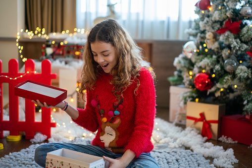 Happy girl opening Christmas present while sitting in front of Christmas tree at home