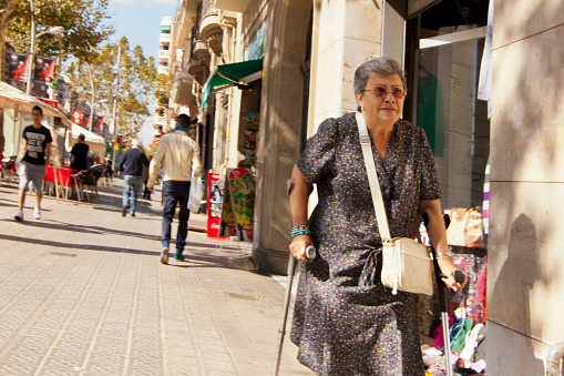 Senior woman walking slowly with walking cane, along the street sidewalk. Barcelona city, Catalonia, Spain.