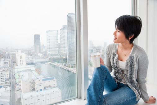 A relaxed young Asian woman, looking out of the window of a modern highrise building. Skyscrapers at Tokyo Bay area in the background. Tokyo, Japan.