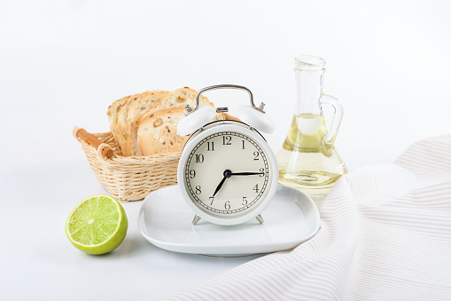 White alarm clock on a white plate against the background of a bottle of oil, bread and lime. It's breakfast time on the alarm clock. Diet concept. Breakfast menu