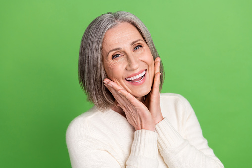 Foto de hermosa señora pensionista de mediana edad las manos se tocan las mejillas riendo disfruta de su nuevo anuncio de crema de loción facial aislado sobre fondo verde photo