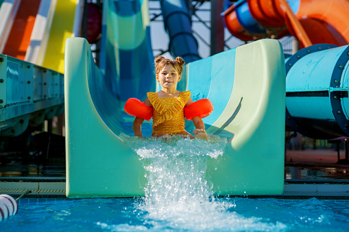 Little preschool girl on water slide in aqua park. Happy child having fun on water slides on family summer vacation in tropical resort. Amusement park with wet playground for young child and baby