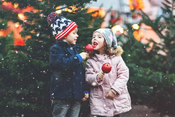 Photo of Two little smiling kids, boy and girl eating crystalized sugared apple on German Christmas market. Happy friends in winter clothes with lights on background. Family, tradition, holiday concept
