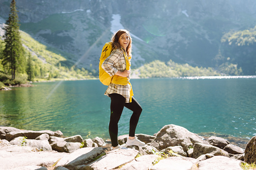 Happy tourist woman with a big yellow backpack enjoys the view of the mountain lake in sunny weather. The concept of hiking, travel, vacation. Active lifestyle.
