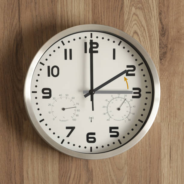 istock A clock, the gray hand points to three o'clock, the black one to two o'clock. The orange arrow indicates the direction of movement of the hands. 1622779496